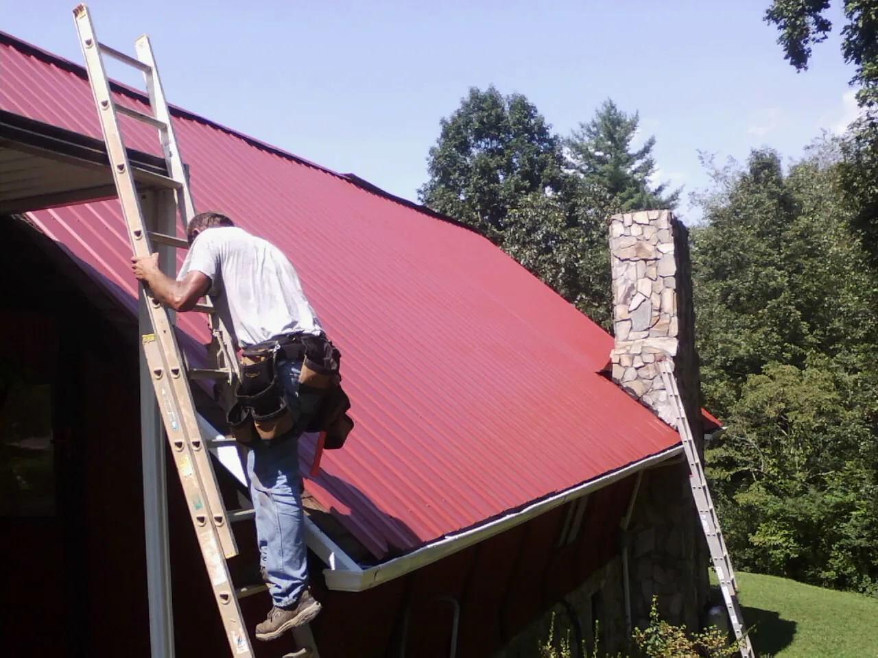 Roofer on a ladder beside a red metal roof and stone chimney — Soffit & Fascia Repair by Roofing Brothers in Hatch